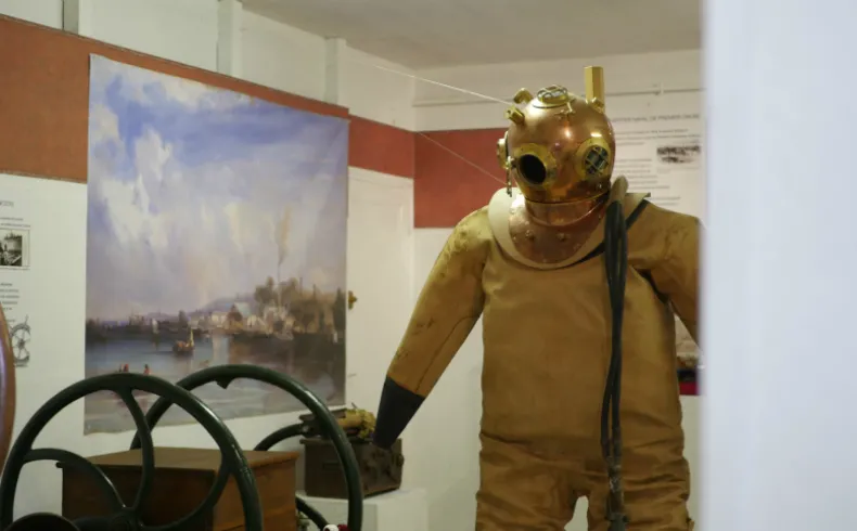 Vintage diving suit mannequin in yellow wetsuit and gilded bronze helmet displayed in a museum, with a ship's wheel and river landscape painting visible behind it.