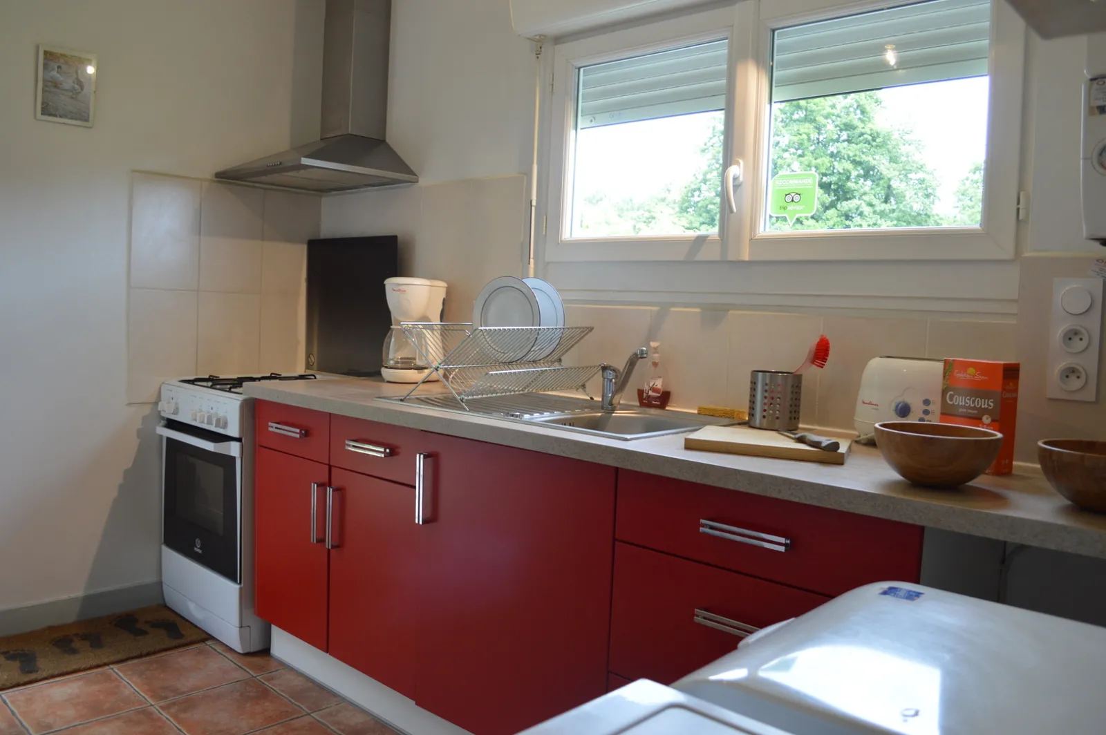 Modern kitchen with red cabinet fronts and grey countertop, equipped with oven, cooker hood, sink and kitchen appliances, with windows overlooking a green garden