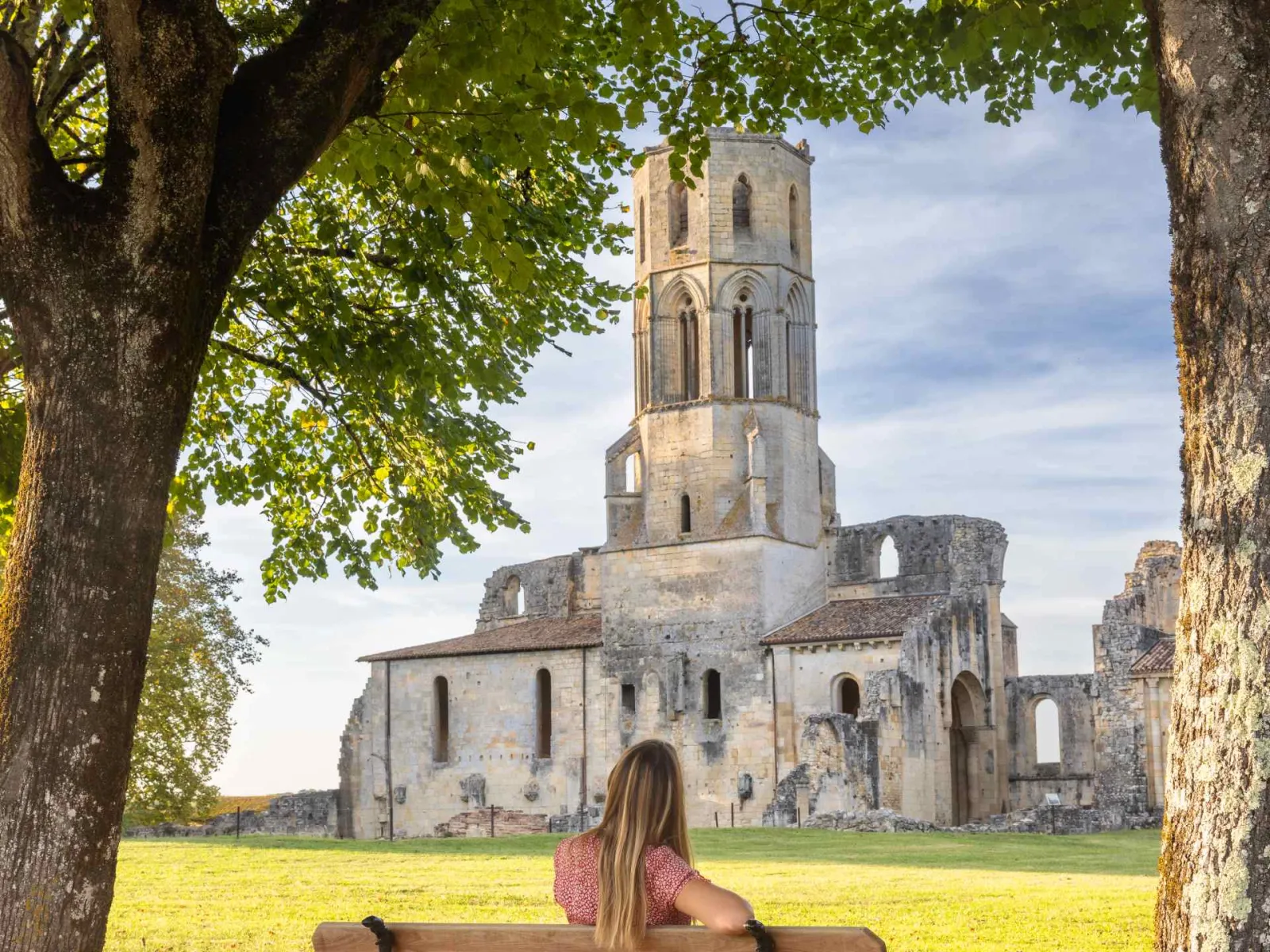 A long-haired woman wearing a pink patterned shirt sits beneath ancient trees facing the ruins of the Abbaye de La Sauve-Majeure near Bordeaux, with its impressive 12th-century bell tower dominating the green landscape.