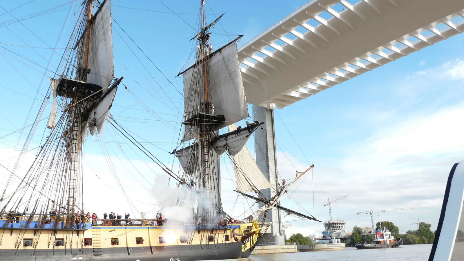 A traditional sailing ship with yellow hull passing under the Jacques Chaban Delmas bridge in Bordeaux, with spectators on deck and construction cranes visible in the background.