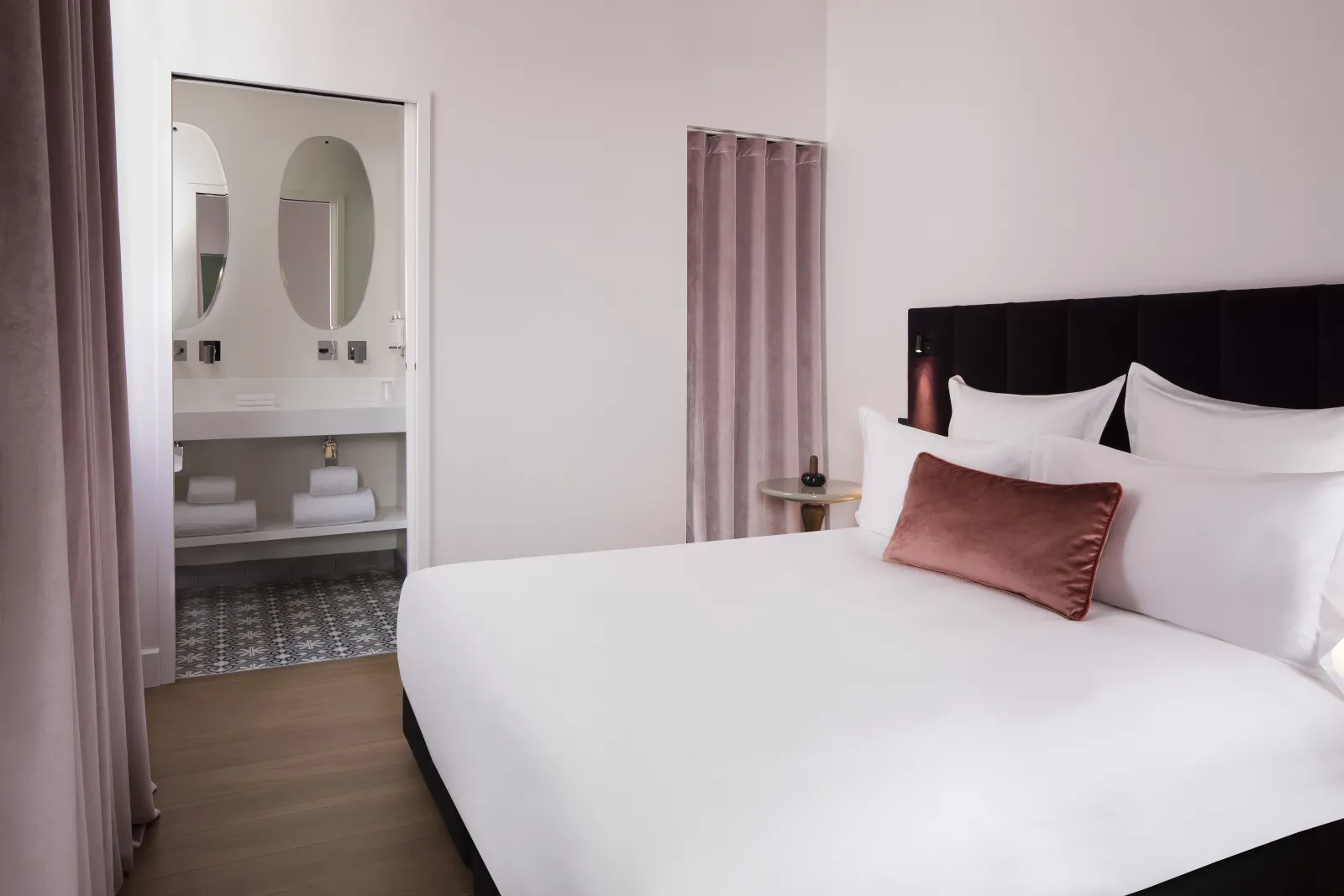 Modern hotel room with white and black bed, open bathroom with oval mirrors, dusty pink curtains and minimalist décor at Hotel Cardinal Bordeaux
