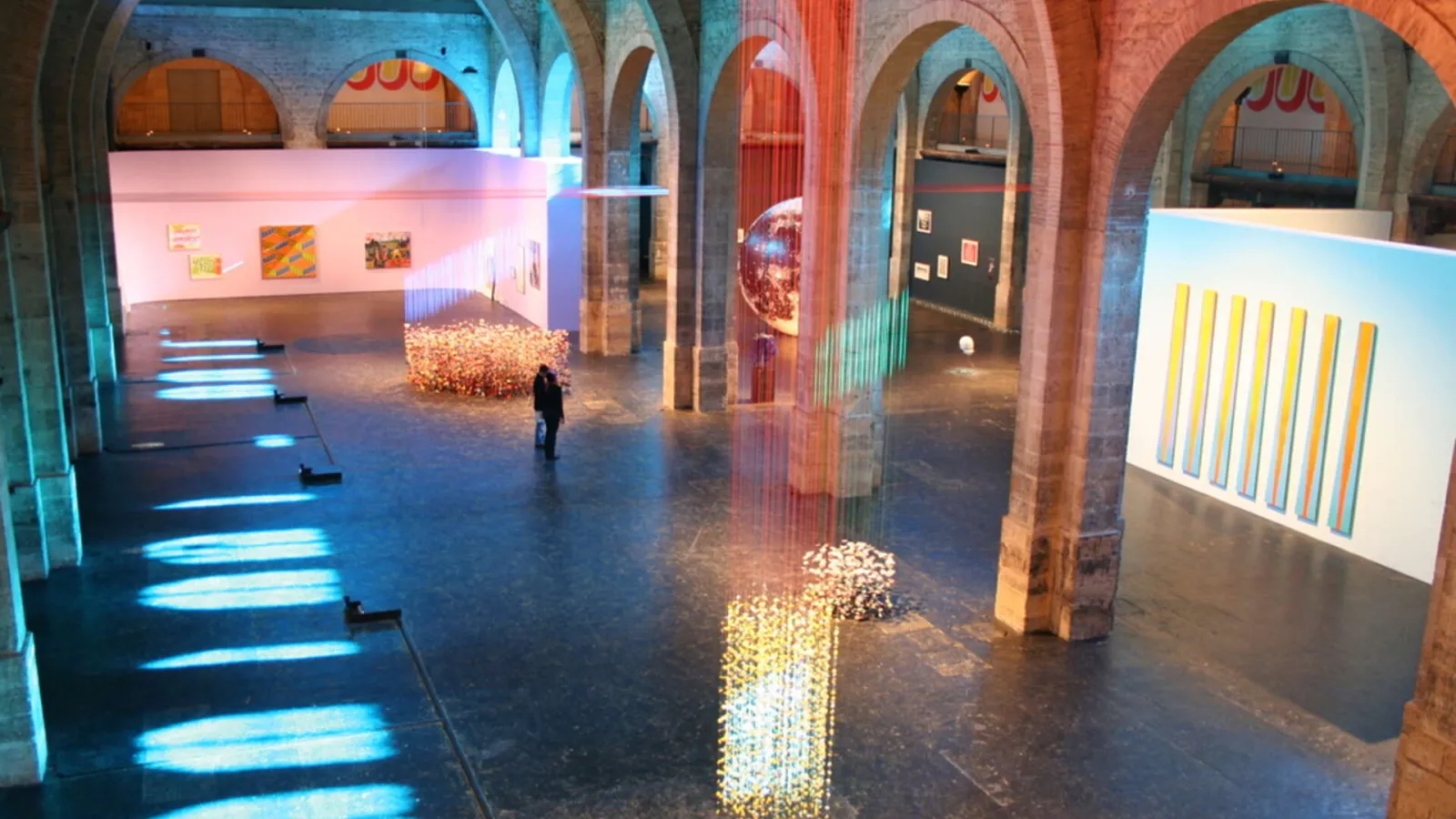 Interior of the CAPC Contemporary Art Museum in Bordeaux featuring a modern art exhibition with luminous installations, red brick columns, art panels, and fluorescent floor elements.