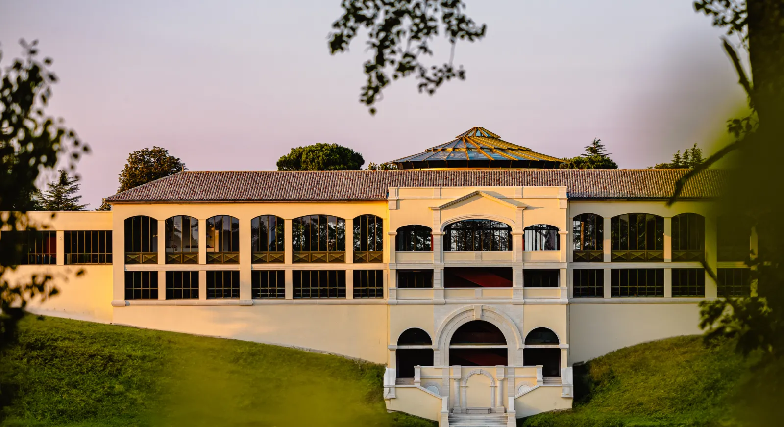 Château Plain Point in Bordeaux, an architectural building featuring yellow and white facade, large bay windows, tiled roof, surrounded by lush greenery and cloudy sky