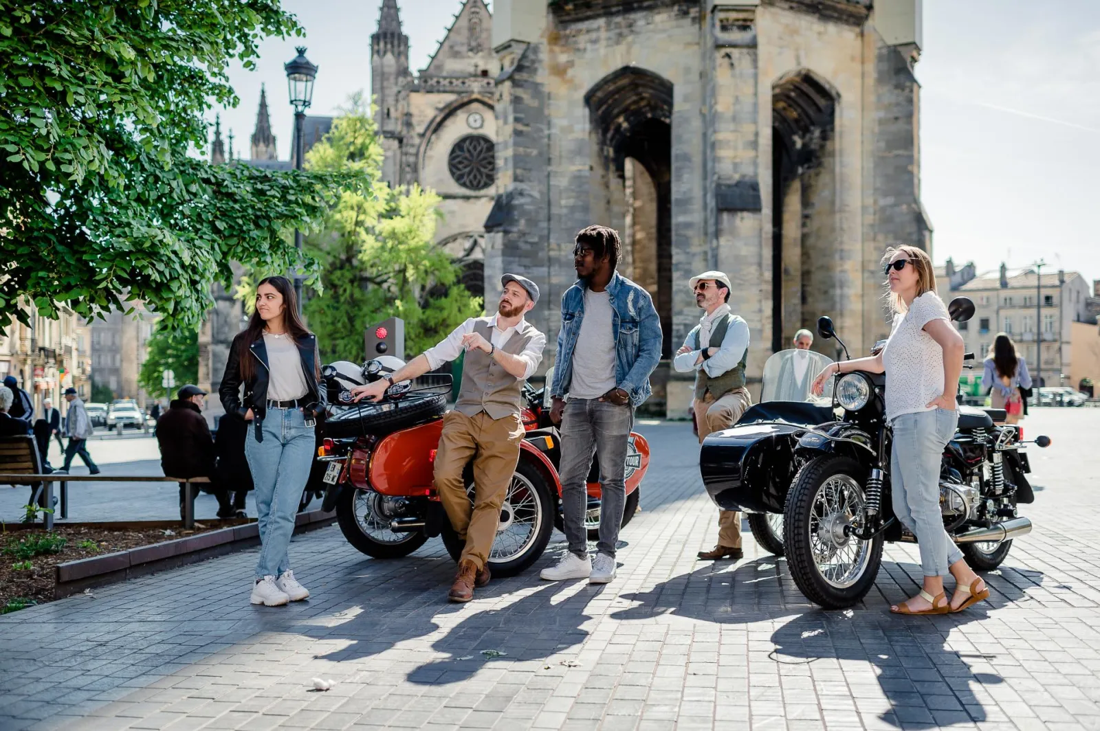 Group of tourists enjoying vintage motorcycles and retro scooters in front of Saint-Michel Basilica in Bordeaux during a themed tour.