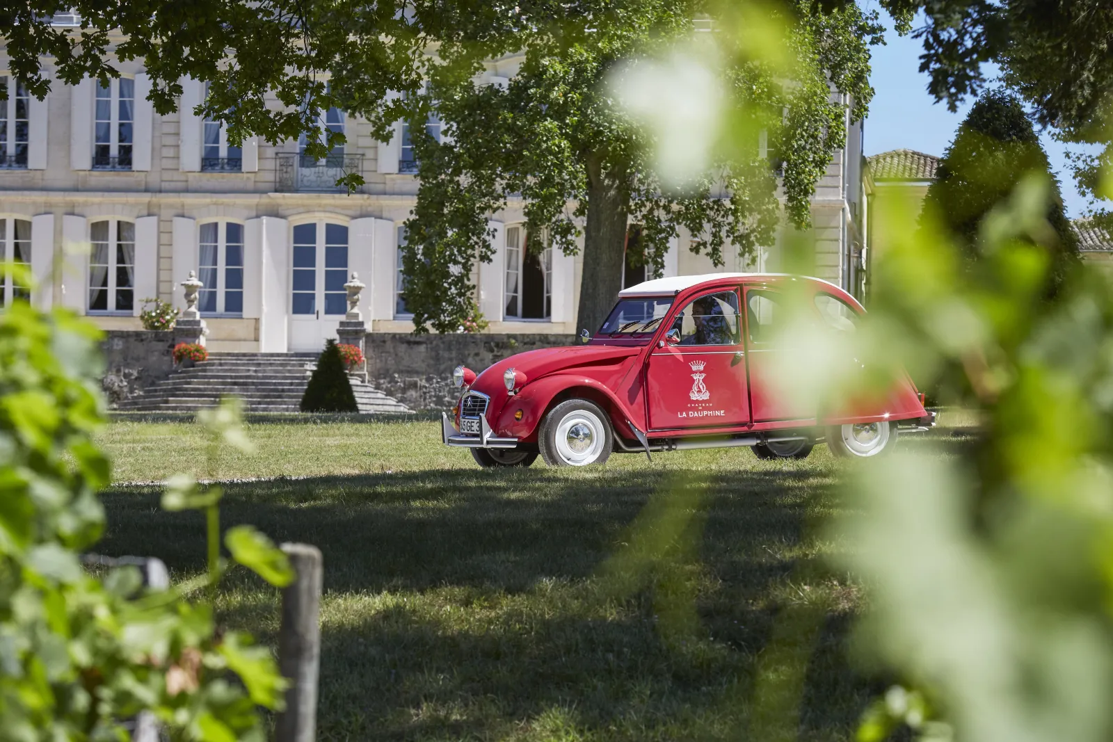 A red Citroën 2CV with « La Dauphine » inscription parked in the grounds of Château de La Dauphine in Bordeaux, with a classical building in the background and trees in the foreground.