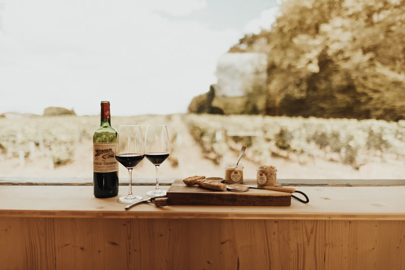 Wine tasting in Saint-Émilion featuring a bottle of Château Chauvin, two glasses of red wine, bread and cheese on a wooden table overlooking the limestone cliffs of Bordeaux.