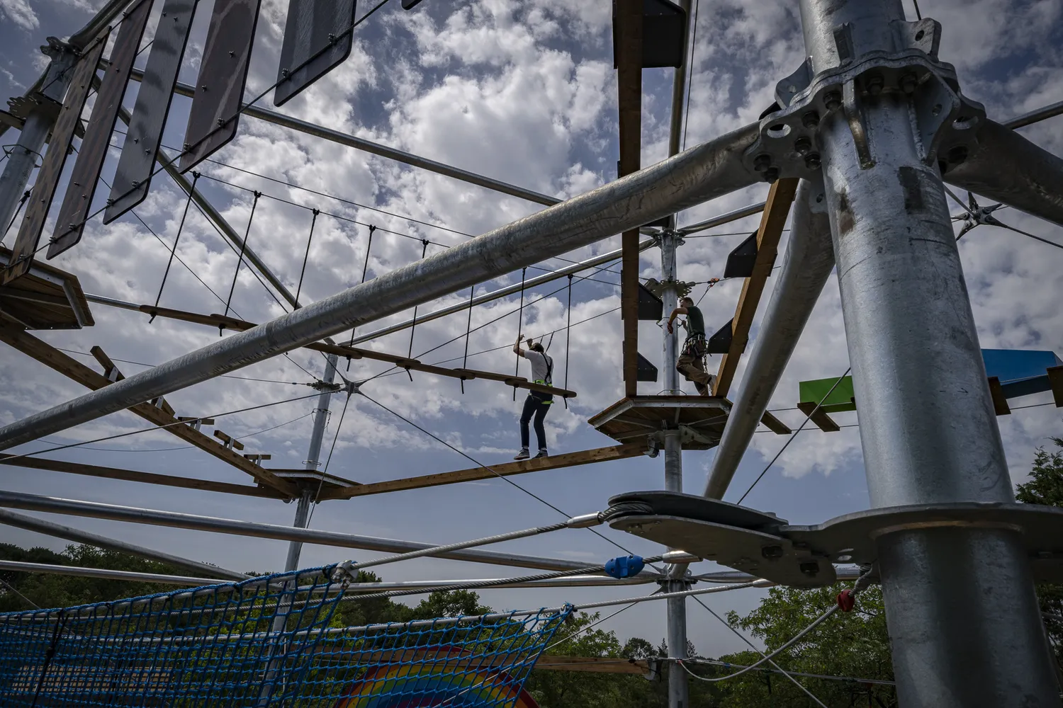 Adventure park with visitors navigating a suspended rope course and beams, featuring metal structures and blue safety nets against a cloudy sky.