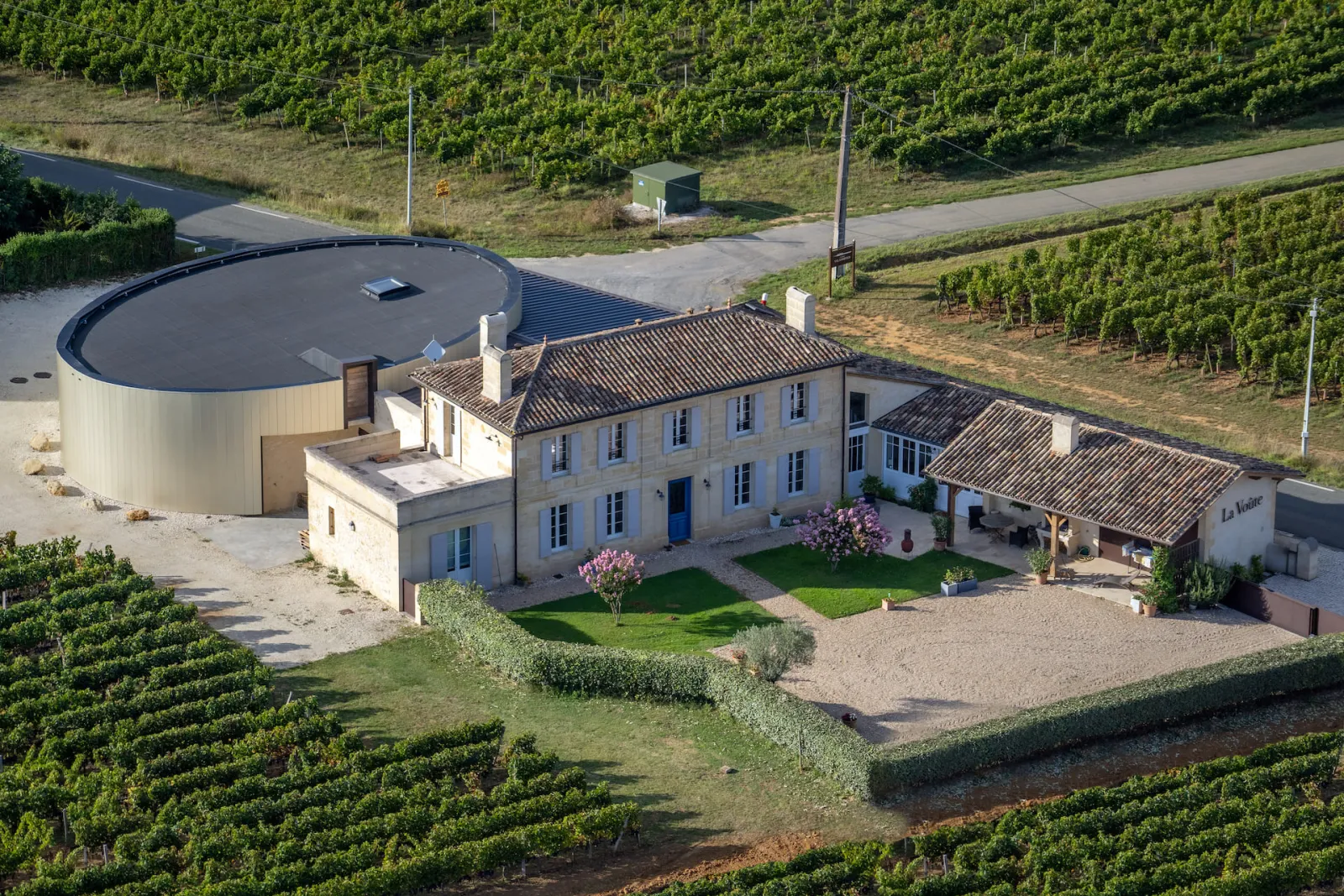 Aerial view of Château La Voûte near Bordeaux, wine estate featuring a stone mansion with tile roof, circular water tank, surrounded by terraced vineyards.