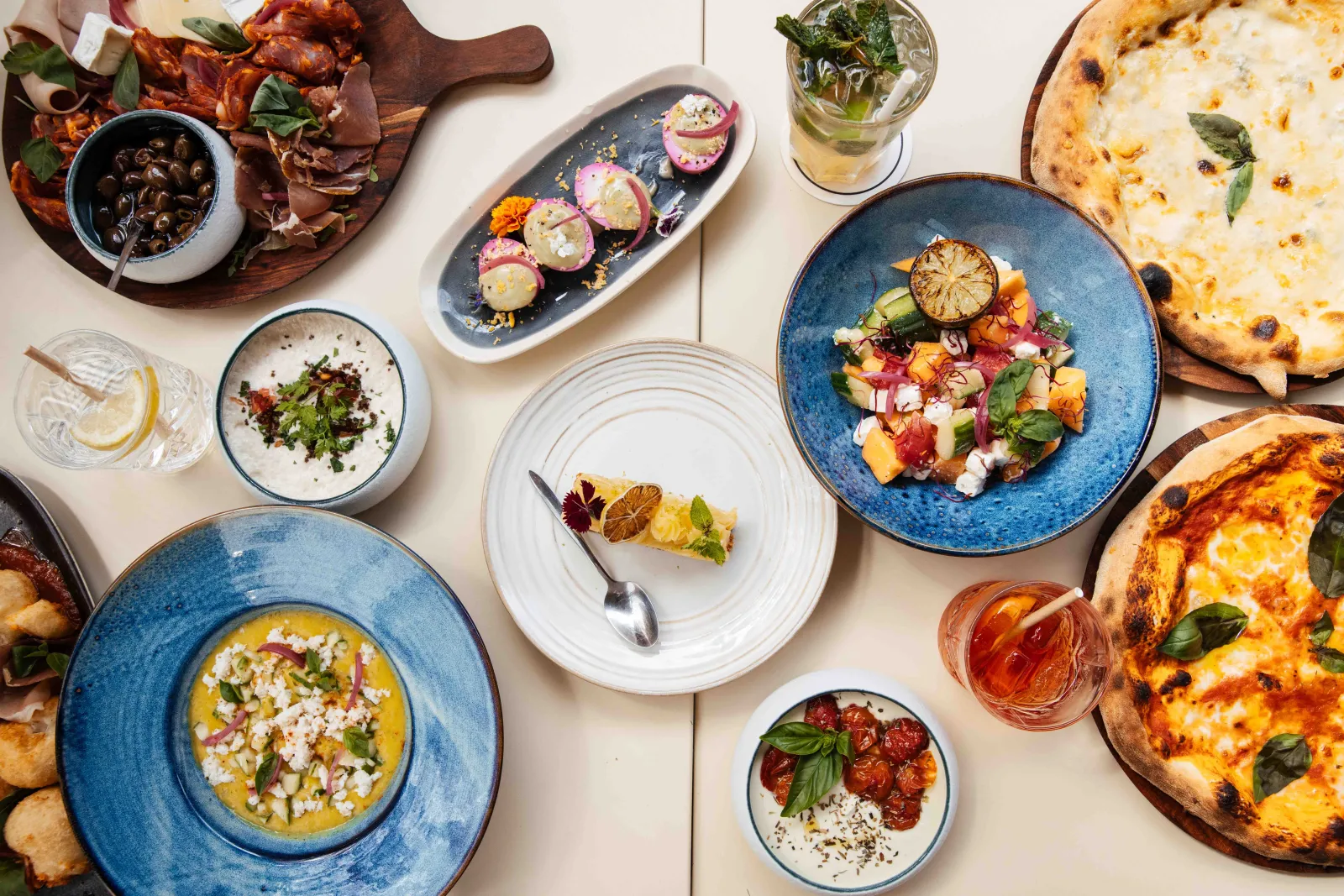 Restaurant table featuring gourmet dishes: wood-fired pizzas, colorful salad, squash soup, cured meats, olives, rosé wine and citrus water glasses, served on blue and white ceramic plates