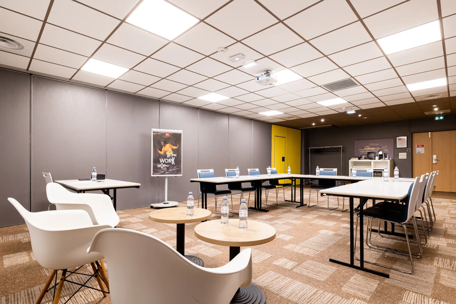 Modern meeting room at Ibis Bordeaux Centre Mériadeck hotel featuring conference tables, white chairs, illuminated panel ceiling, gray walls with yellow accents, water bottles, and motivational poster.