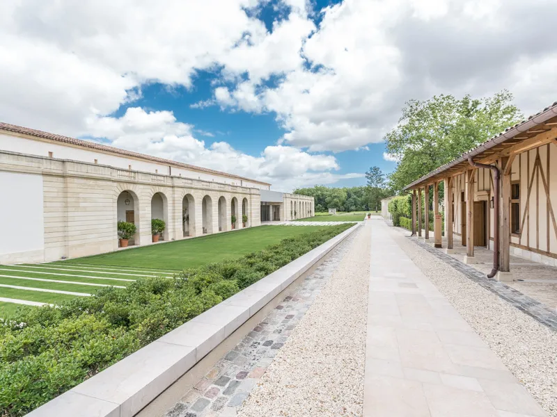 Central pathway of a Bordeaux château featuring white colonnaded building on the left, timber-framed structure on the right, manicured lawn and cloudy blue sky