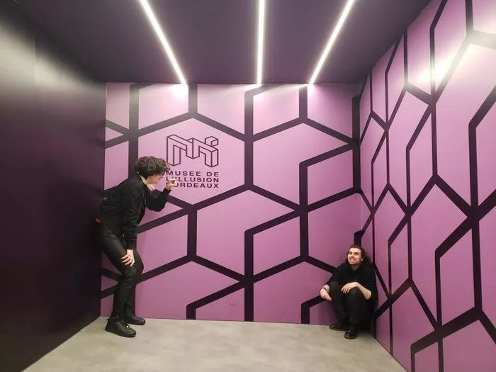 Two visitors in a room at the Museum of Illusion in Bordeaux, surrounded by pink walls with black geometric patterns and white neon lights on the ceiling.