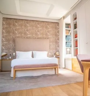 Elegant hotel bedroom featuring a double bed with white linens, textured beige headboard, light wood furniture, and built-in shelving in a bright, contemporary setting.