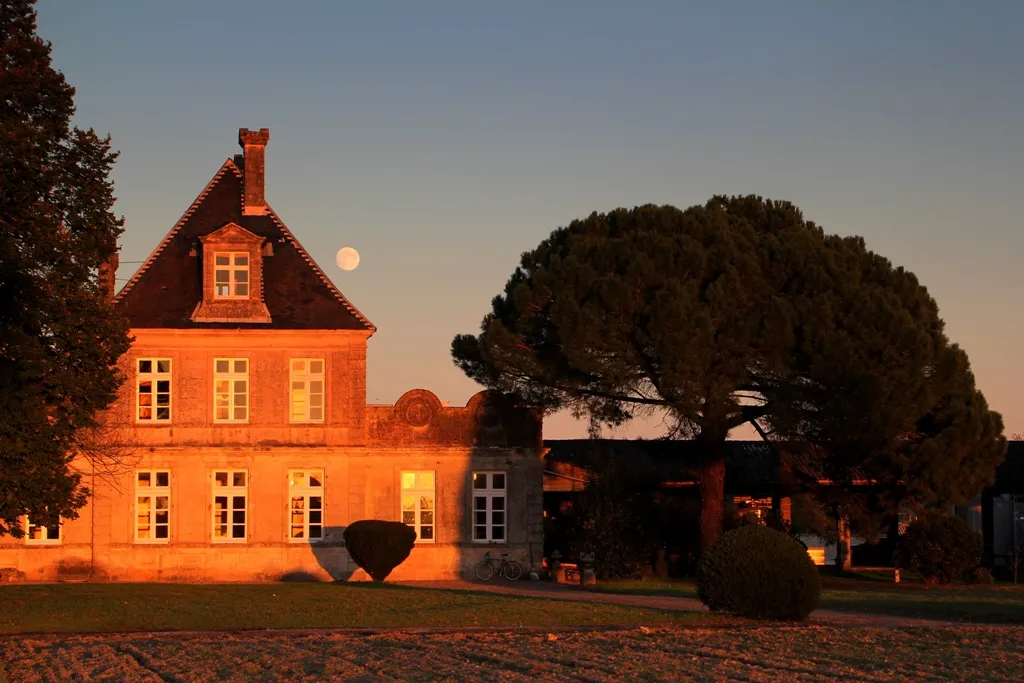 Château de Cérons in Bordeaux glowing in golden sunset light, featuring bright orange walls, mansard roof, and illuminated windows, surrounded by landscaped gardens and trees.