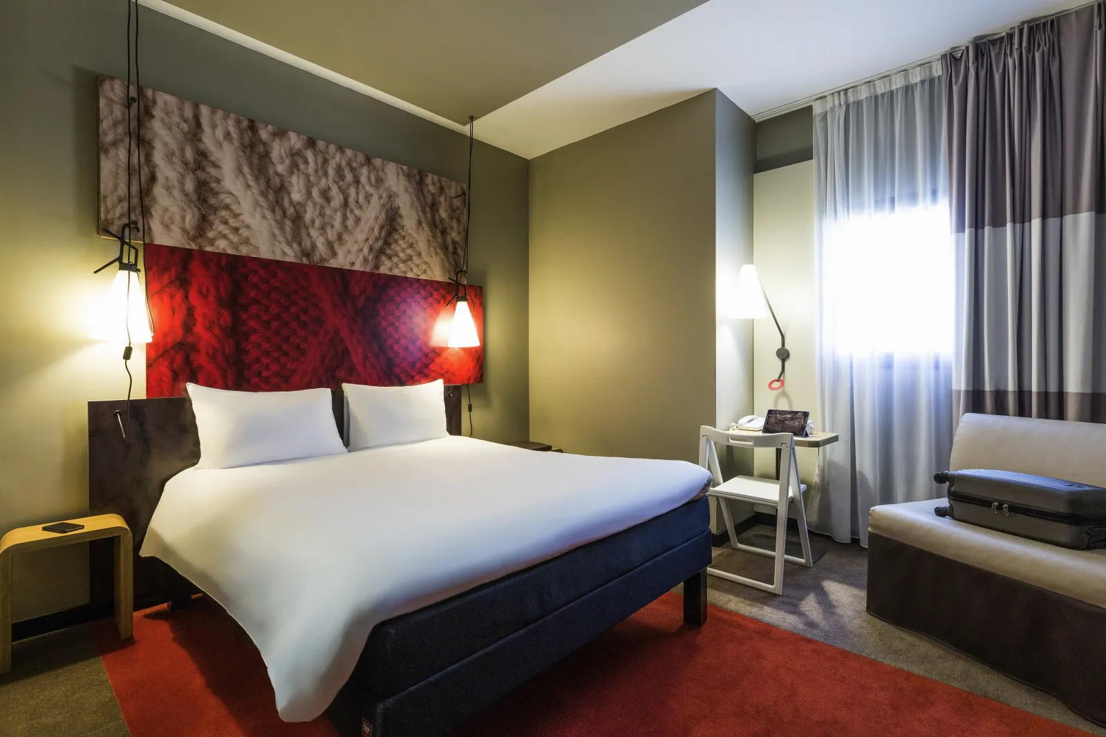 Modern hotel room featuring a double bed with white linens, red and brown headboard, pale yellow walls, warm ambient lighting, and a window with gray curtains at Ibis Bordeaux Centre Gare Saint-Jean.