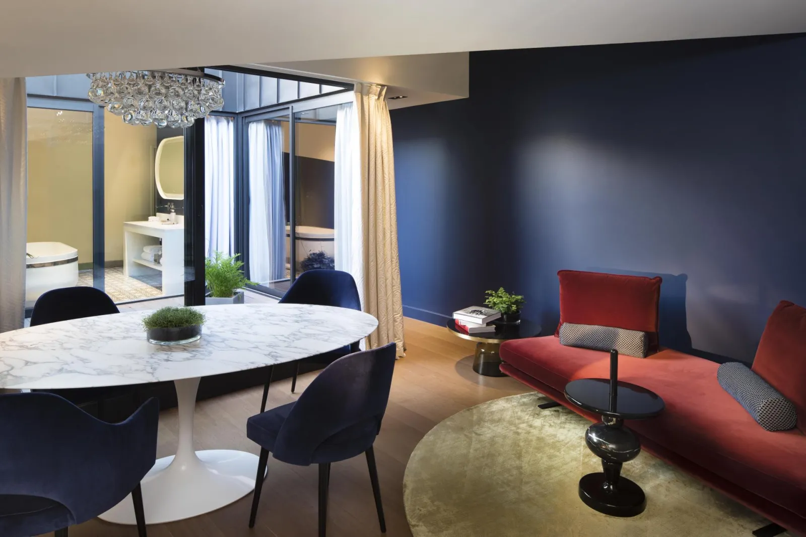 Luxury suite at Hotel Cardinal Bordeaux featuring a modern living space with red sofa, white marble dining table, black chairs, a spa-like bathroom with walk-in shower, and navy and gold tones