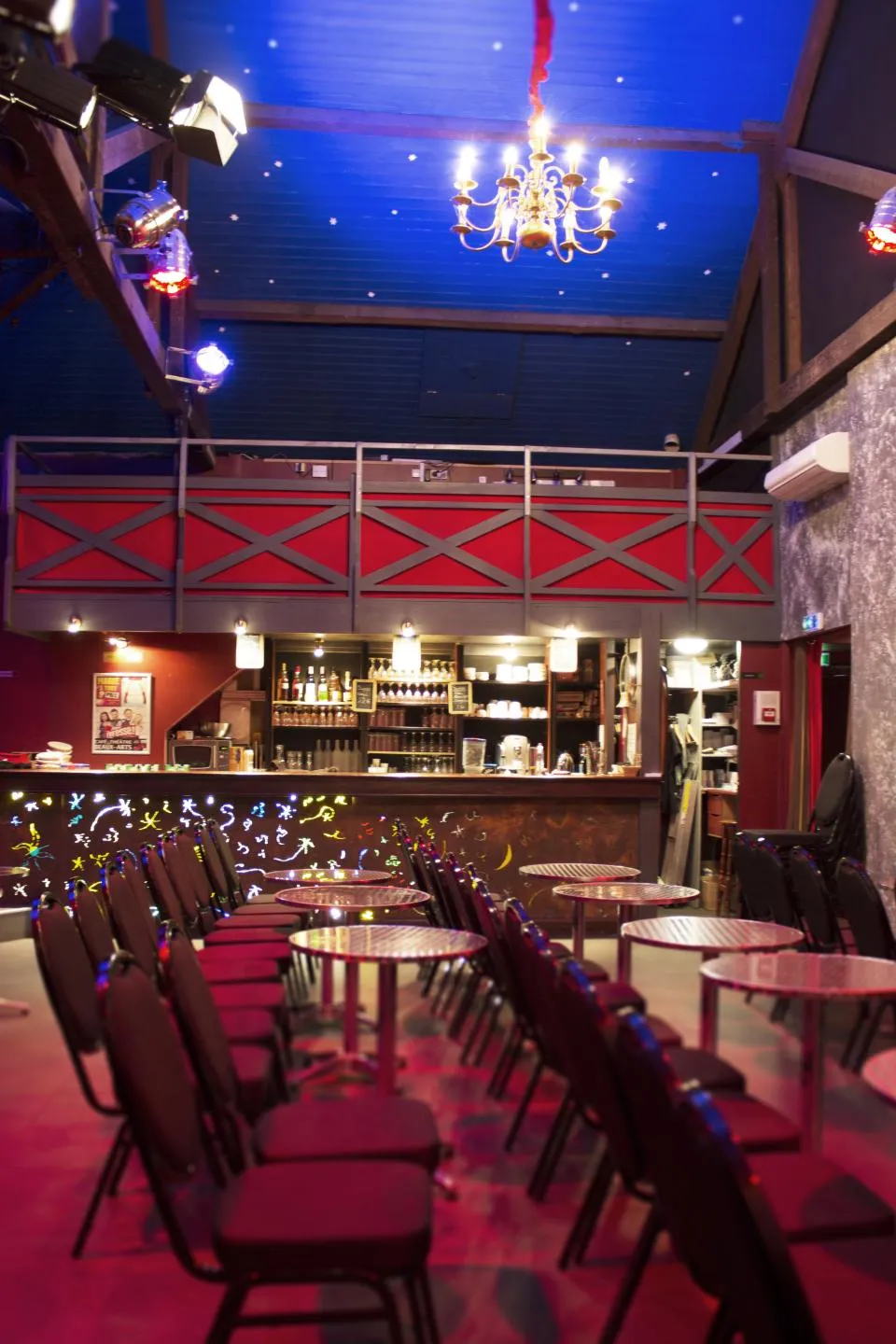 Interior of Bordeaux's Beaux Arts Theater featuring an elegant hall with burgundy walls, starlit blue ceiling, golden chandelier, bar in the background, and round tables with black chairs.