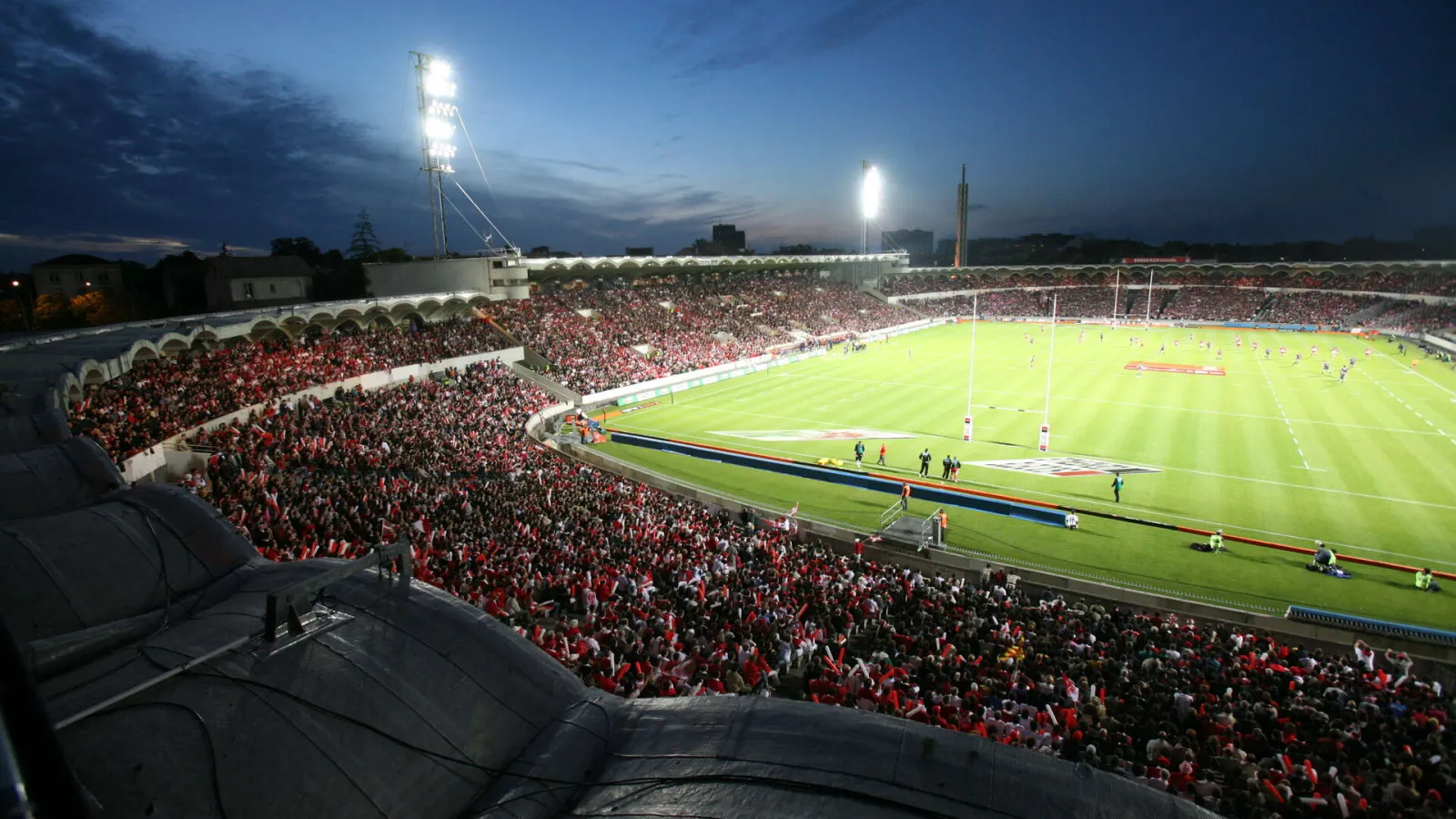 Aerial view of the illuminated Chaban-Delmas Stadium in Bordeaux during evening, filled with spectators in red and white attire, rugby pitch in the foreground with crowded stands.