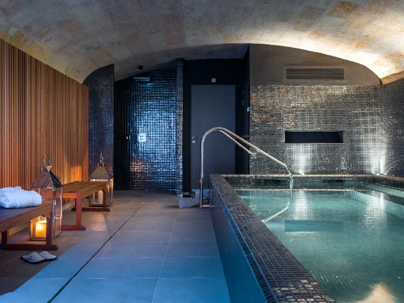 Underground indoor swimming pool at Villas Foch hotel in Bordeaux, featuring a turquoise tiled basin, access stairs, relaxation area with loungers and candles, beneath a raw concrete vault.