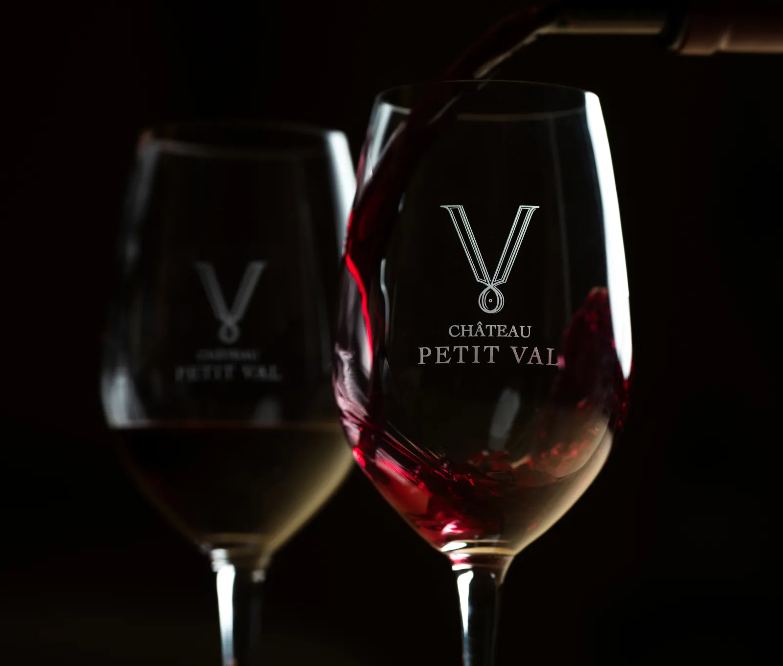 Two engraved wine glasses with Château Petit Val logo filled with red and white wine against a dark background, showcasing Bordeaux wines.