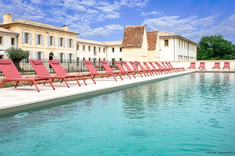 Swimming pool at Château Fage in Bordeaux featuring red loungers along turquoise water, with white stone buildings and terracotta roofs in the background