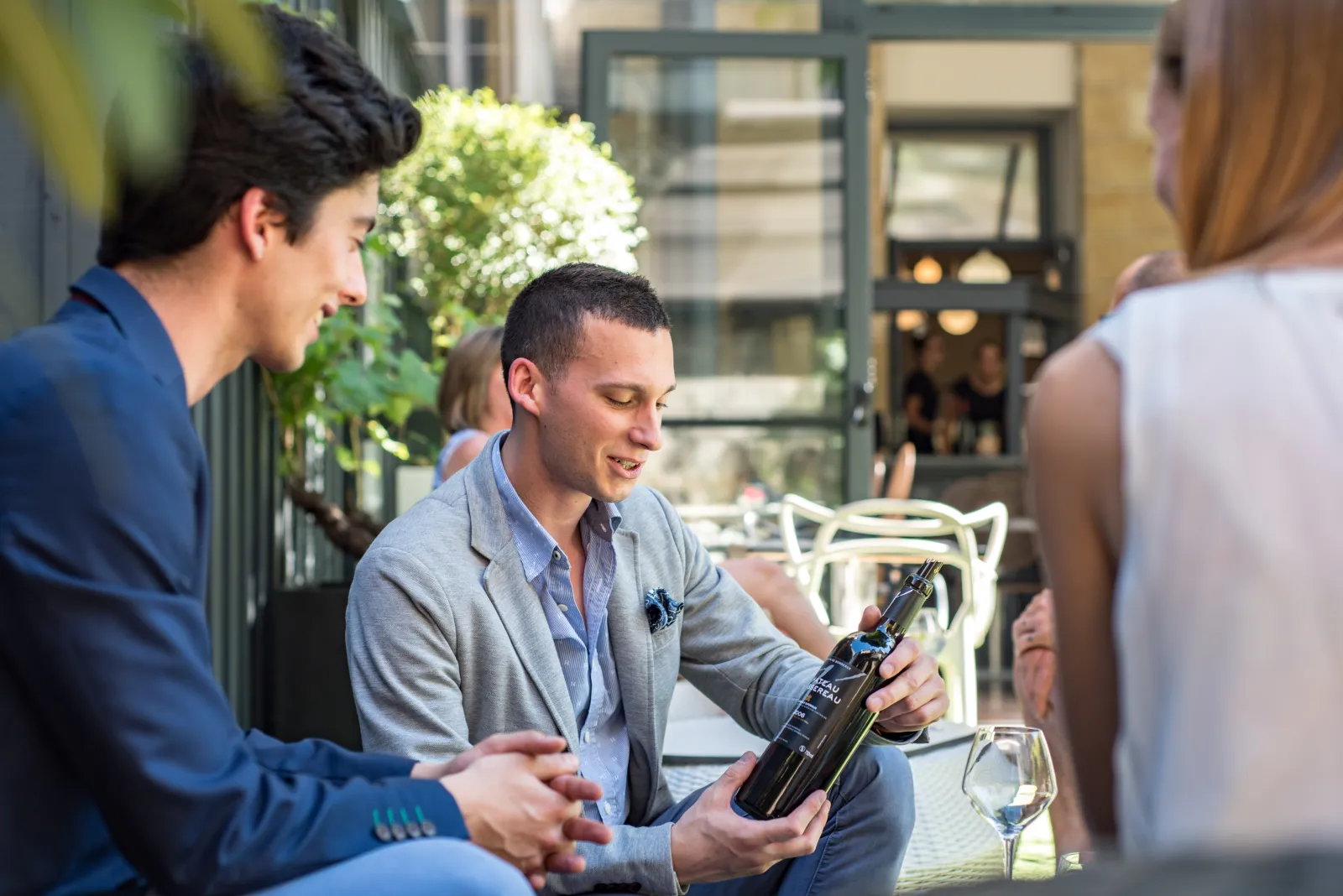 A man in a gray shirt presents a wine bottle to customers in the courtyard of a boutique hotel in Bordeaux, with green plants and wine glasses in the background.