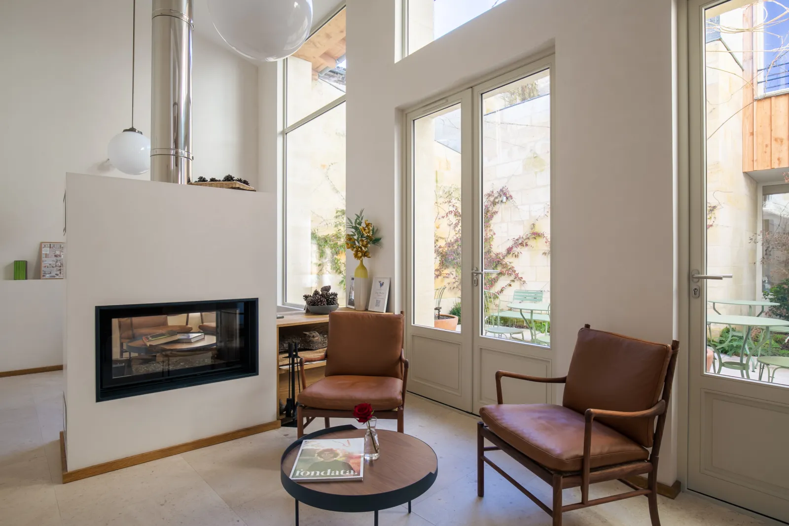 Bright living room at Badon Boutique Hotel in Bordeaux featuring two brown leather armchairs, a modern fireplace, round coffee table, floor-to-ceiling glass doors opening onto a patio, and contemporary minimalist décor.
