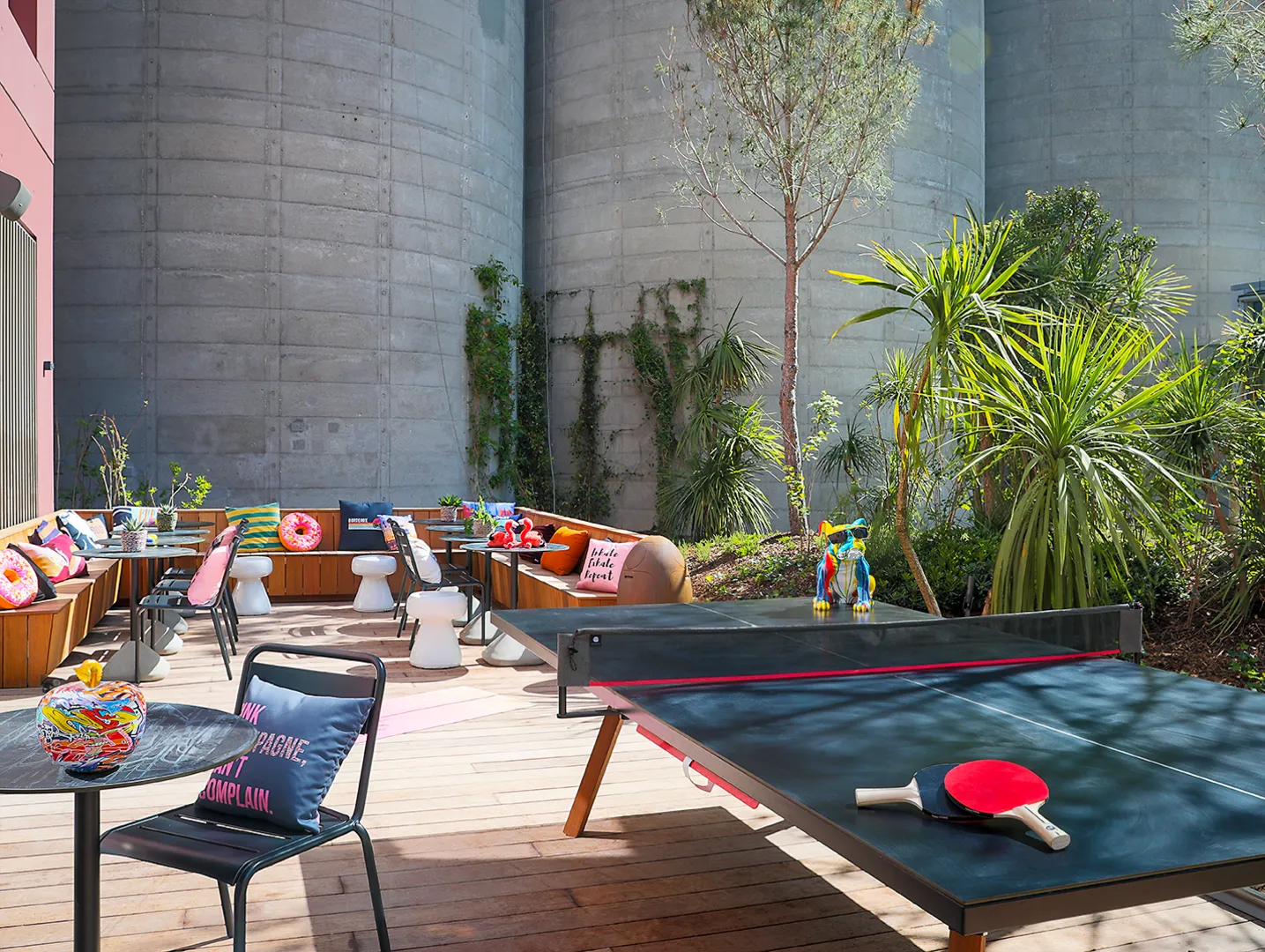 Colorful outdoor terrace at Moxy Bordeaux featuring a ping-pong table, wooden furniture, tropical plants, and concrete walls