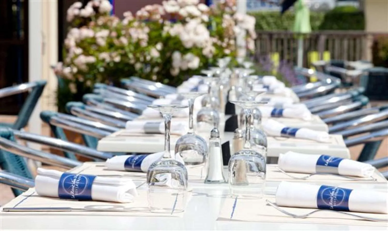Elegantly set conference table with white linens featuring blue stripes, crystal glasses, blue chairs, and white flowers in the background at a Bordeaux garden venue.