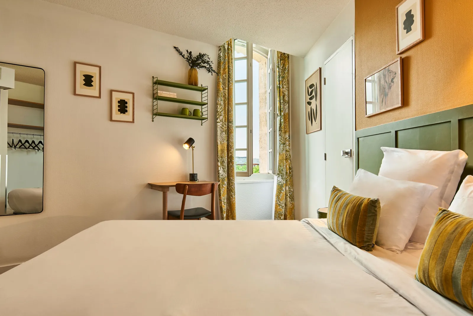 Modern and elegant hotel bedroom featuring a queen-size bed with green headboard, yellow striped cushions, tall window with gold curtains, wooden desk, round mirror, green wall shelves and contemporary décor in Bordeaux