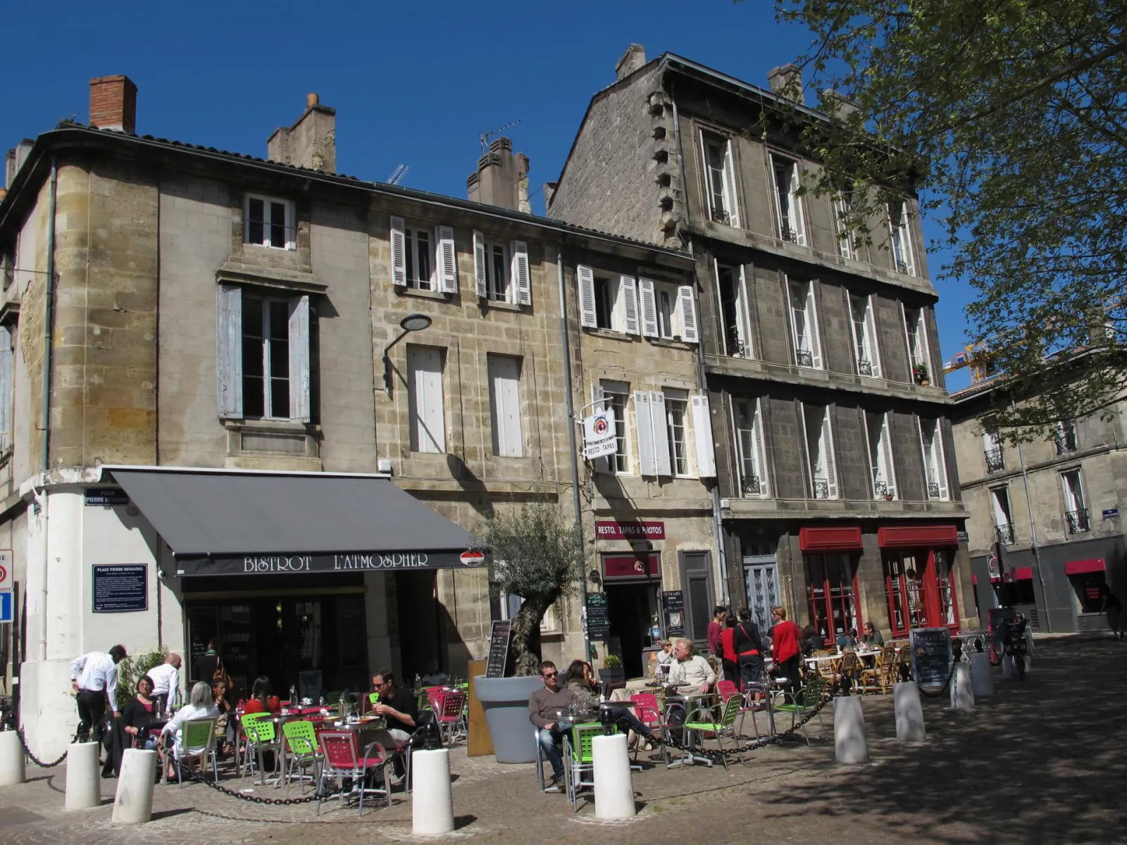 Outdoor café terrace with colorful chairs in front of Haussmann-style stone buildings in Bordeaux, with the Abbatiale Sainte-Croix visible in the background under clear skies.