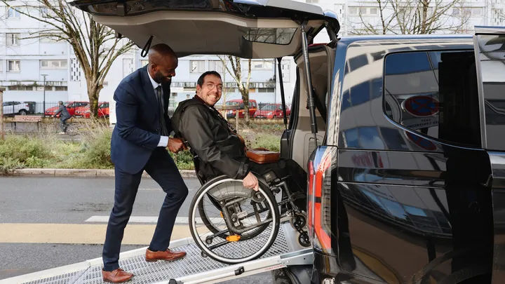 A man in a blue suit assists a person in a wheelchair boarding a minibus equipped with an access ramp in Bordeaux. The vehicle features an adapted transportation system for people with reduced mobility.