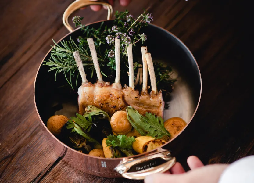Rack of lamb with exposed bones garnished with fresh herbs and roasted vegetables in a serving dish