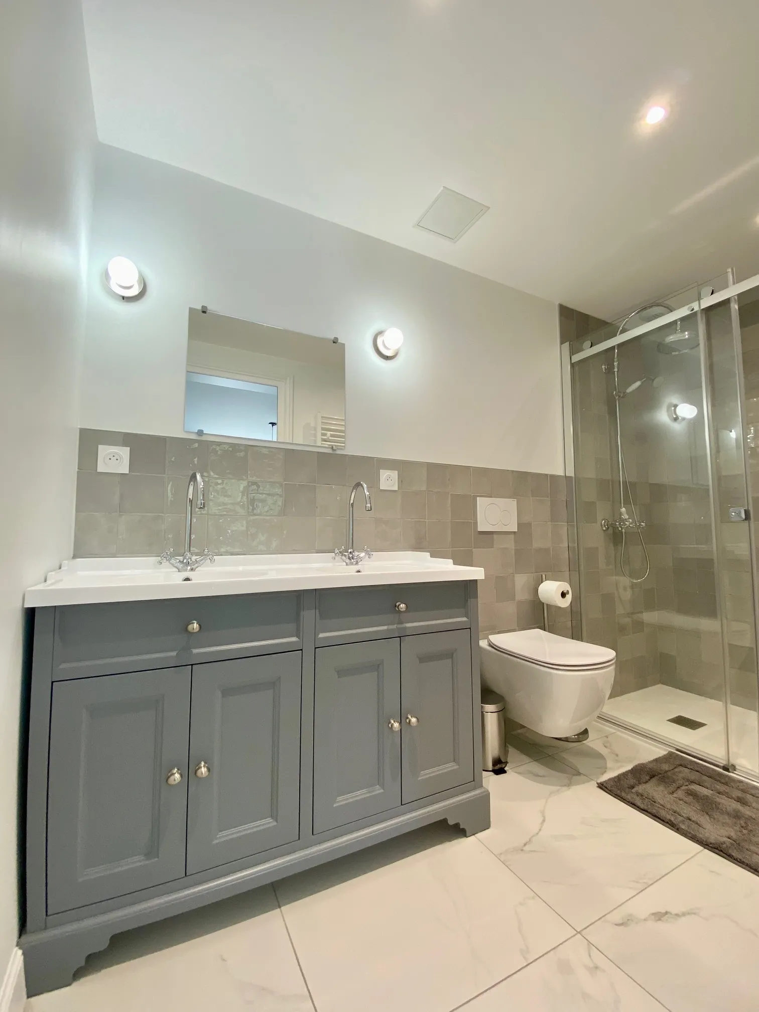Modern bathroom featuring a dual-sink vanity with dark gray cabinetry, illuminated mirror, neutral tile work in beige and gray tones, walk-in shower with glass enclosure, and wall-mounted toilet.