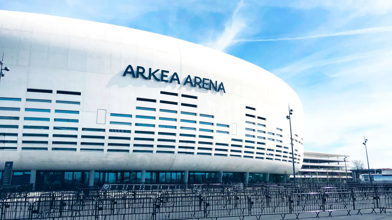 Facade of Arkea Arena in Floirac, Bordeaux, featuring its distinctive white structure with blue and black louvers against a clear blue sky.