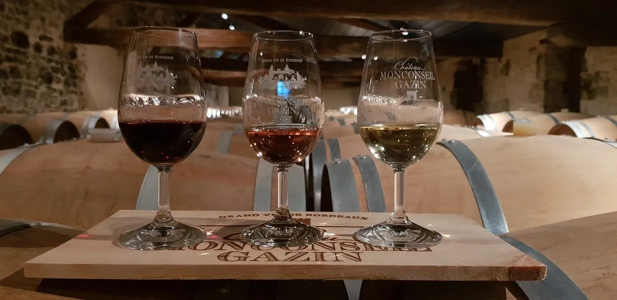 Three wine tasting glasses from Château Monconseil-Gazin containing Bordeaux wines of different colors, arranged on a wooden table in a cellar with barrels in the background
