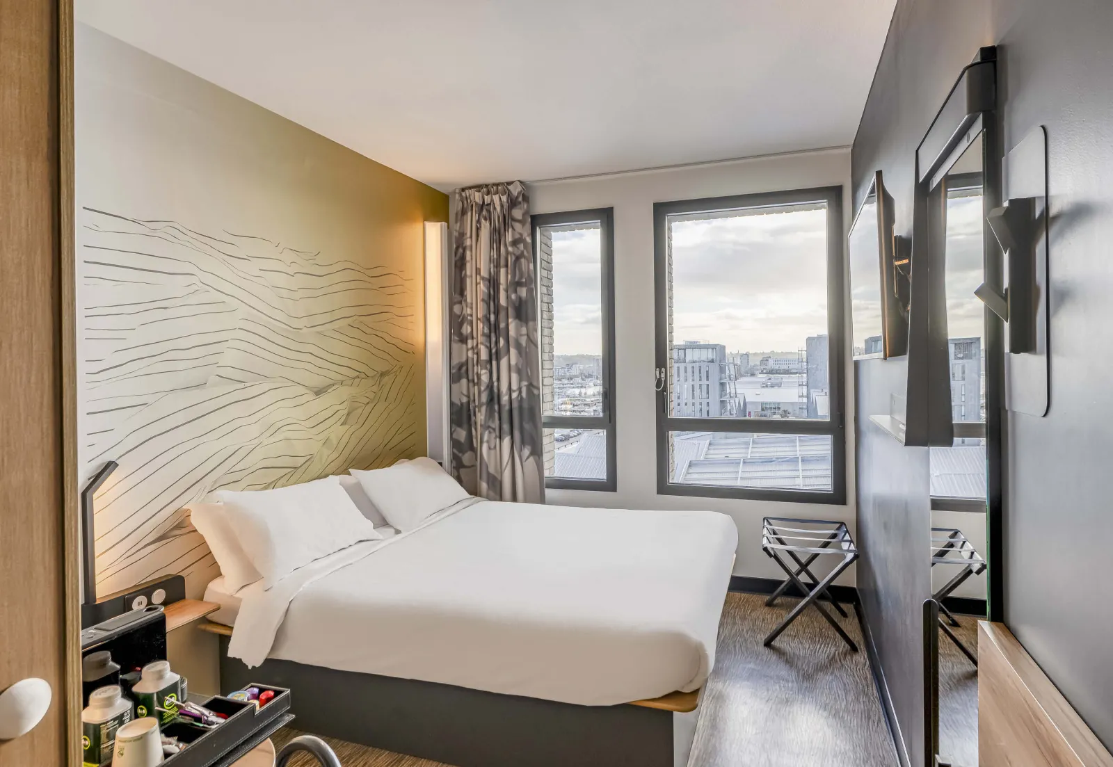 Modern hotel room with double bed with white linens, yellow headboard with wavy patterns, large windows overlooking Bordeaux cityscape, contemporary furnishings and view of the Bassins-à-Flot.