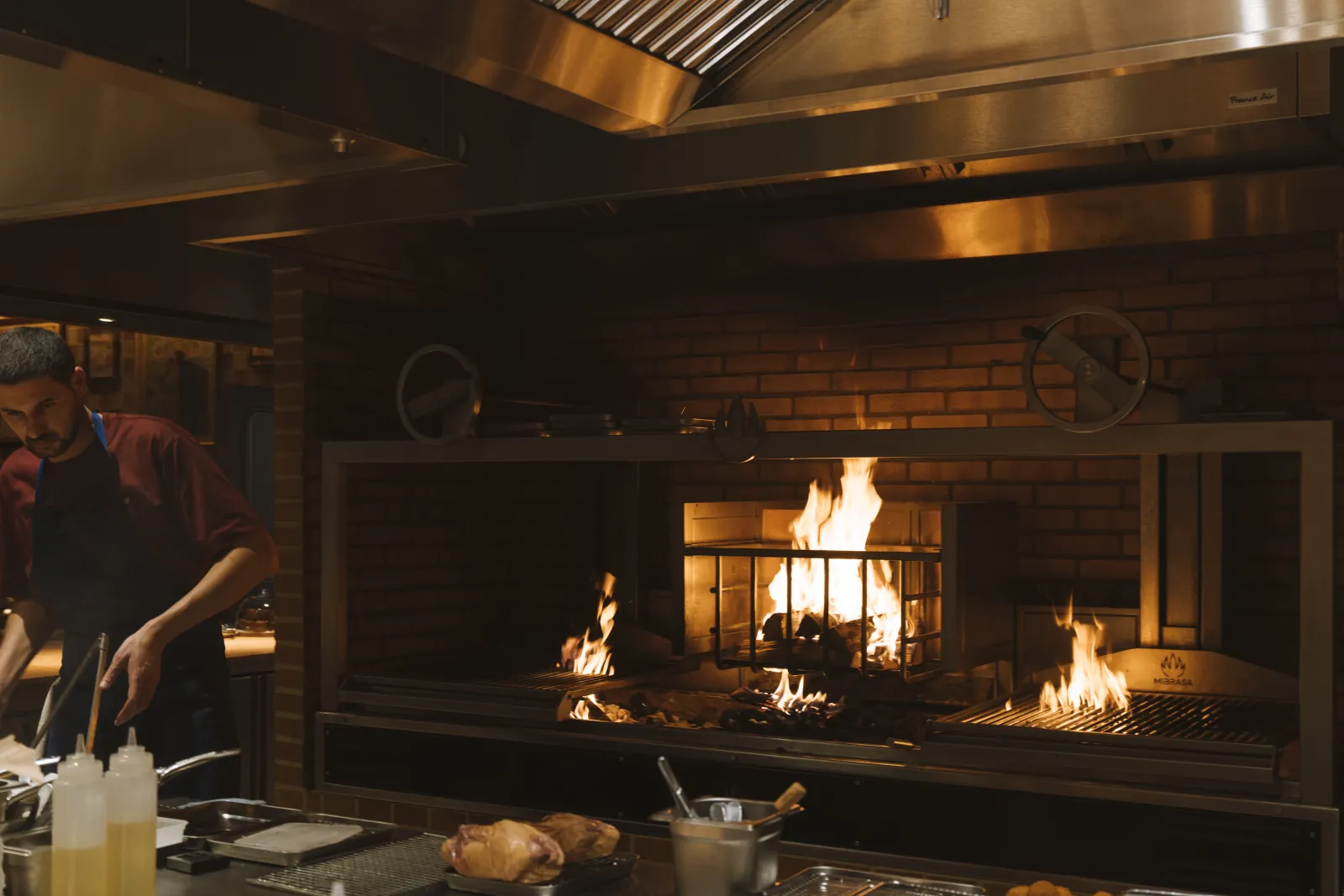 Modern brasserie interior featuring a large central fireplace with steel and red brick, active flames, open kitchen counter with food preparations, client in background, warm ambient lighting