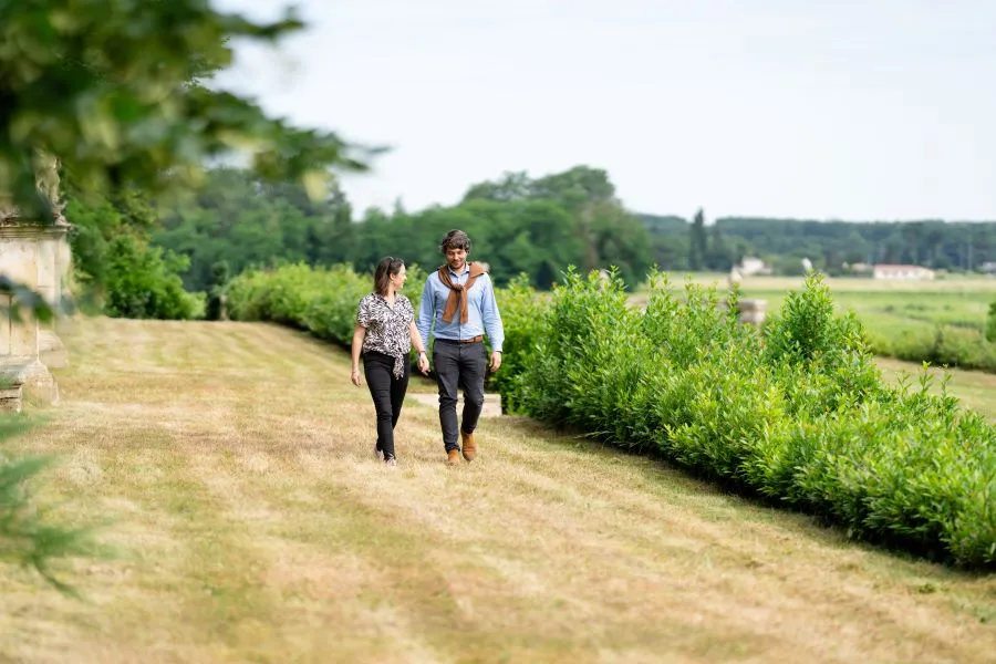 Two people walking on a dirt path alongside a green hedge in the vineyards of Château de Malle near Bordeaux, with fields and trees in the background.