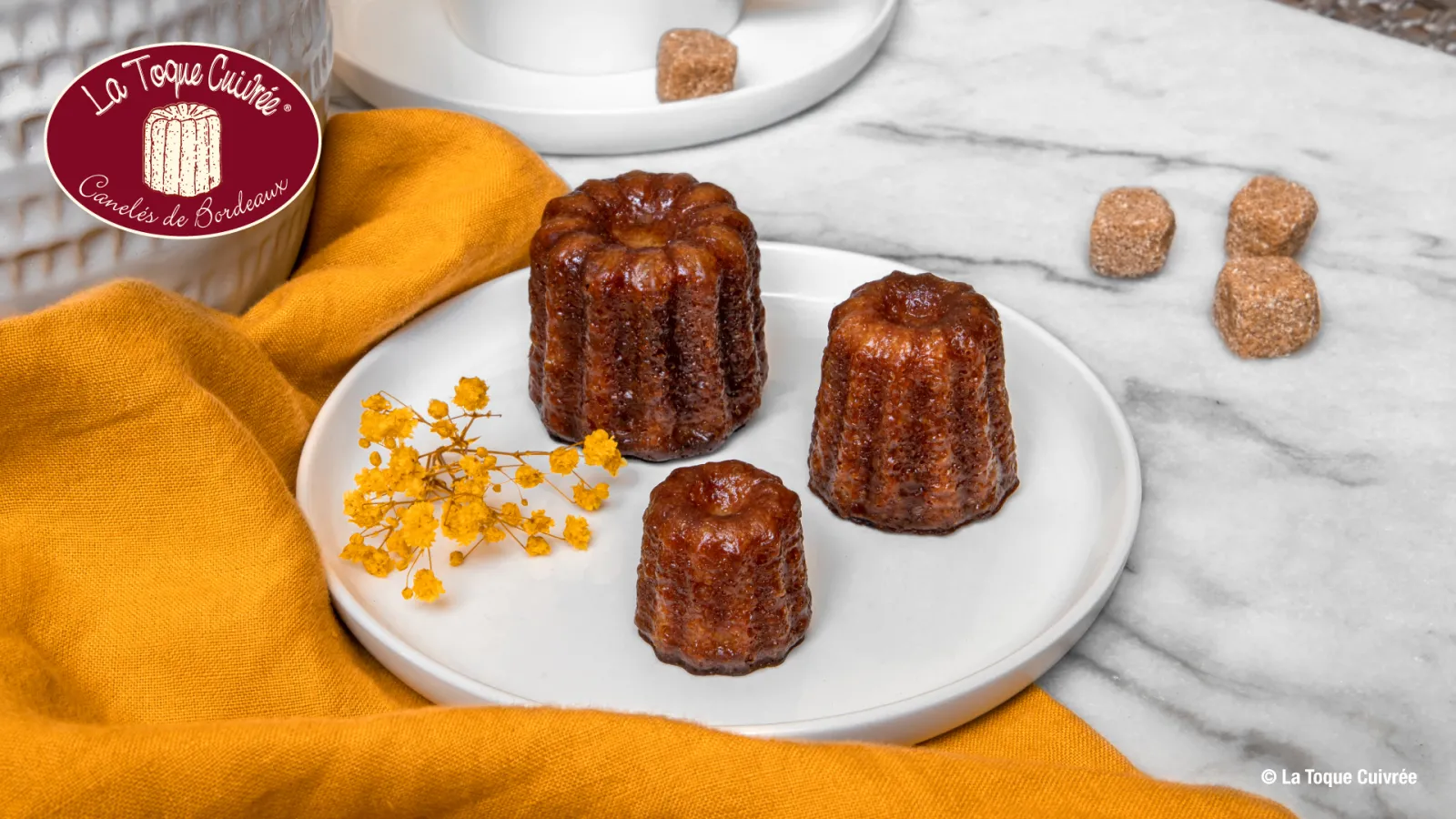 Three golden and shiny Bordeaux canelés presented on a white plate, decorated with yellow dried flowers, with orange fabric and crystallized sugars in the background