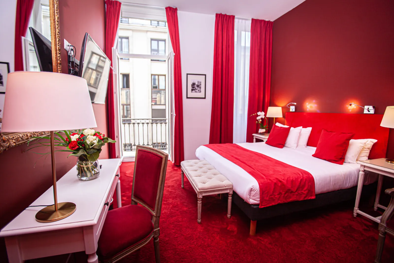 Luxury hotel bedroom in red and pink tones featuring a king-size bed, gold floor lamp, red curtains, balcony with Bordeaux view, and elegant décor.