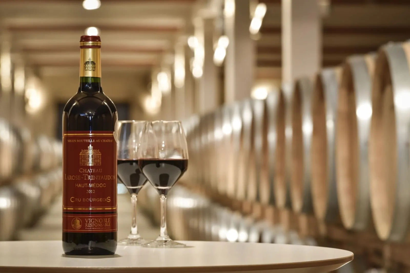 Bottle of Château Larose-Trintaudon Haut-Médoc 2012 red wine with two filled glasses displayed in a Bordeaux winery cellar lined with oak barrels.