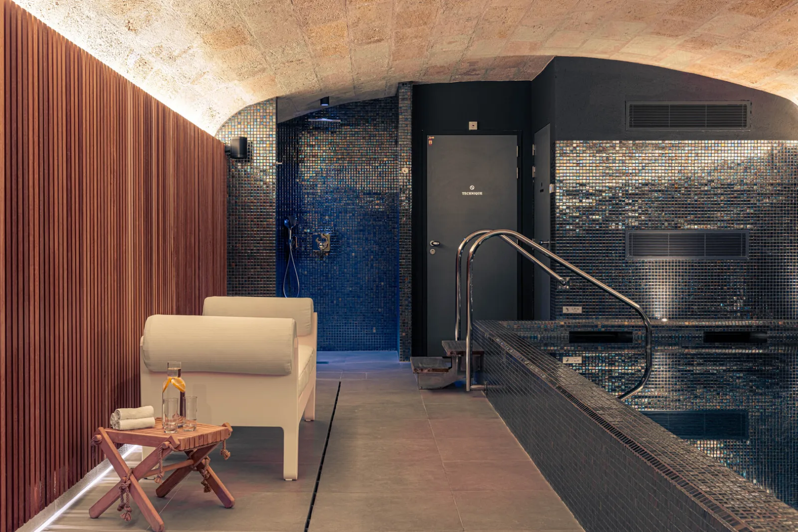 Luxury spa featuring a blue tiled pool, rainfall shower, sauna, beige relaxation armchair and wooden furnishings at Hôtel Villas Foch in Bordeaux