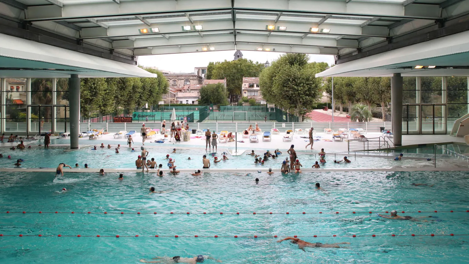 Municipal swimming pool Judaïque Jean Boiteux in Bordeaux featuring indoor pools with retractable roof, crowds of swimmers and sunbathers, exterior patio terrace, and historic buildings visible in the background