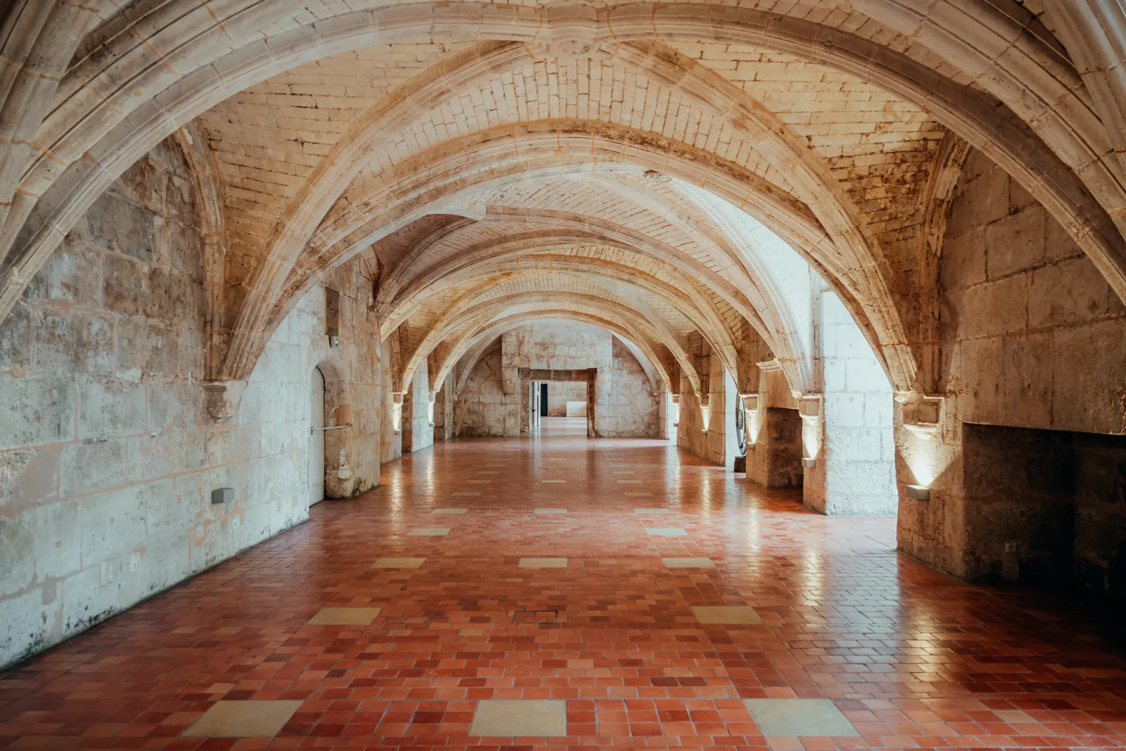 Vaulted corridor with white and beige brick arches, stone columns, and red and beige tiled flooring creating a receding perspective in a historic castle.