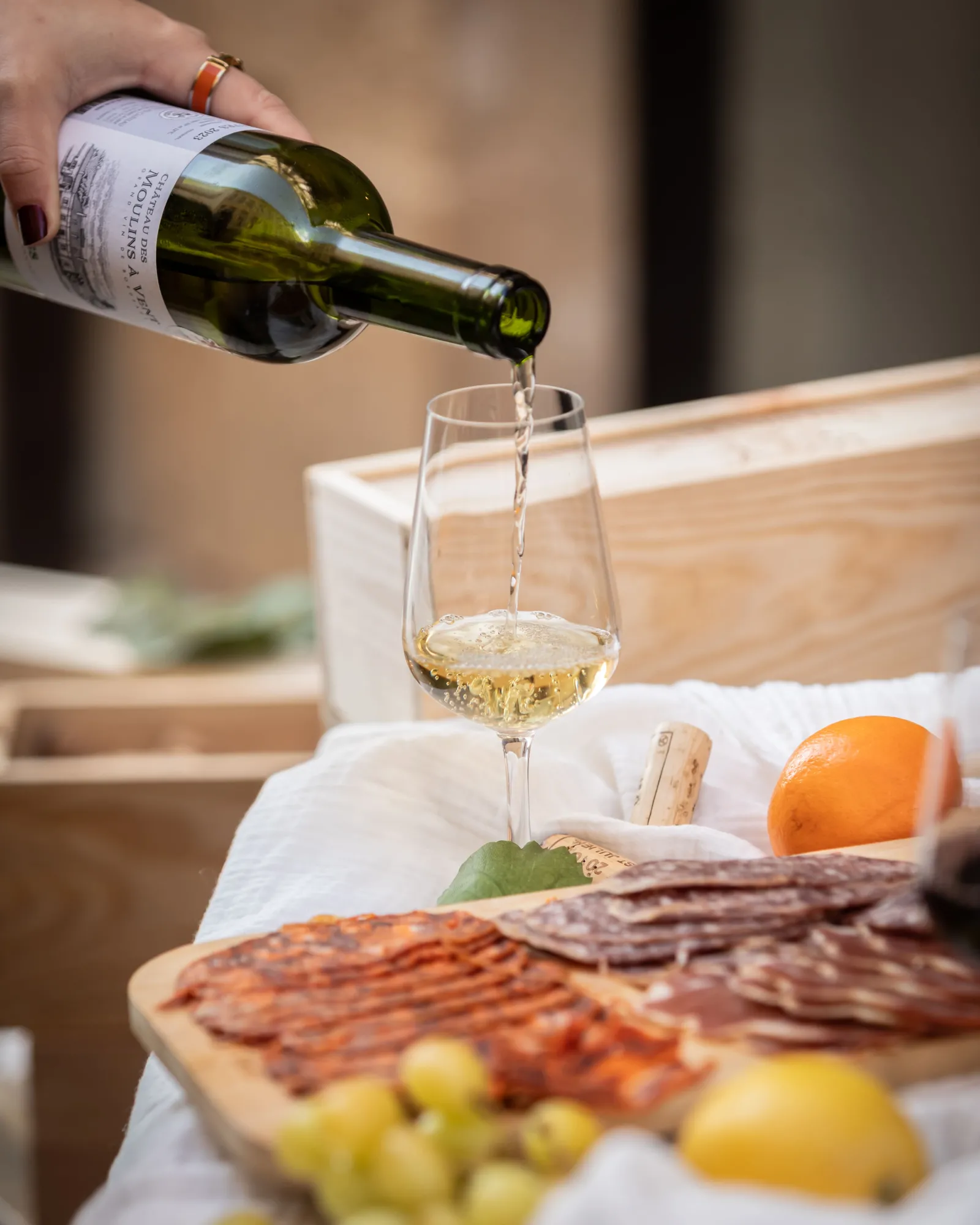 Pouring a glass of dry white Bordeaux wine into a wine glass, accompanied by a charcuterie board with cured meats, dried fruits and olives on a wooden table.