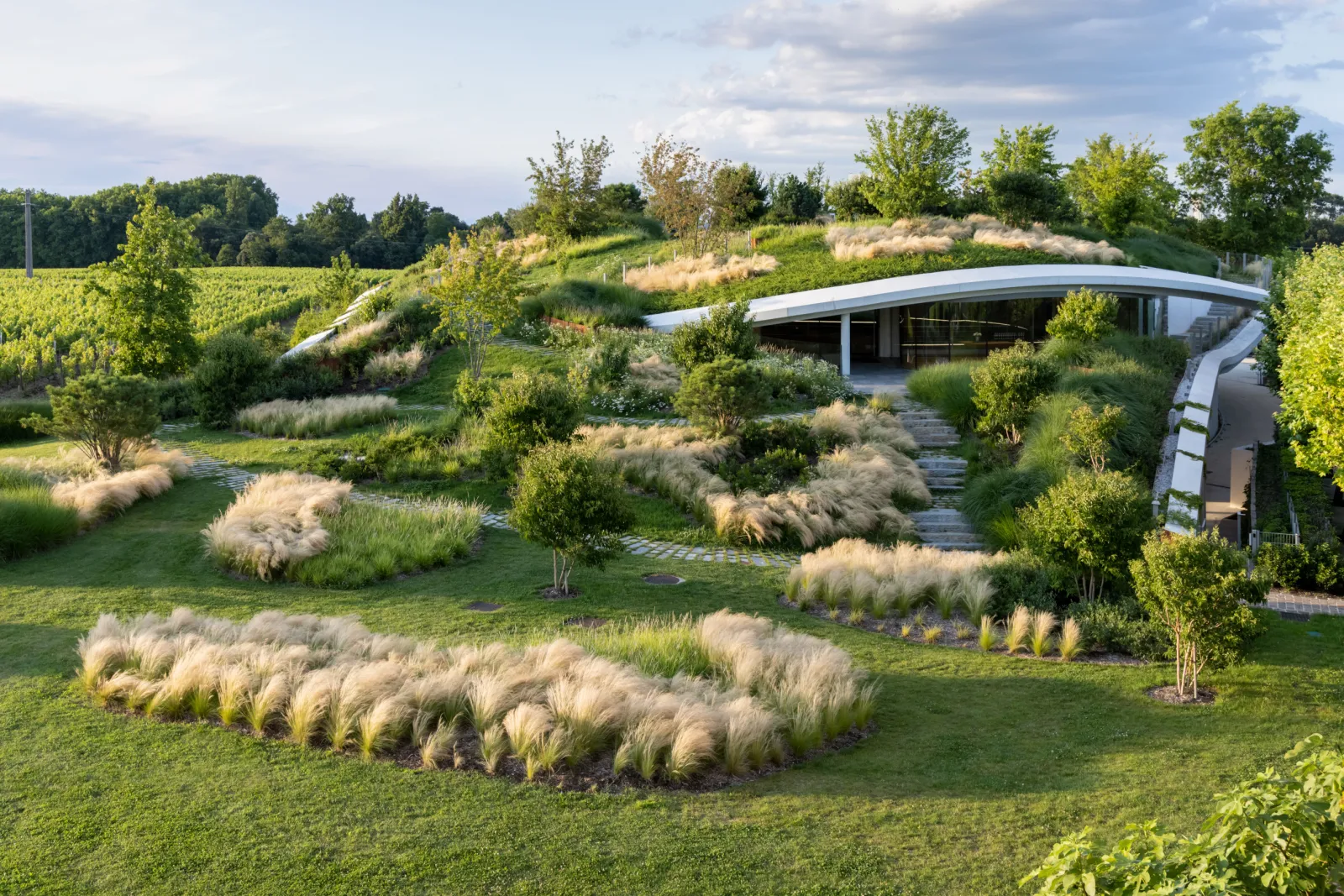 Aerial view of Château Haut-Bailly near Bordeaux featuring contemporary curved architecture, surrounded by landscaped gardens with ornamental grasses, fruit trees, and water features overlooking vineyards.