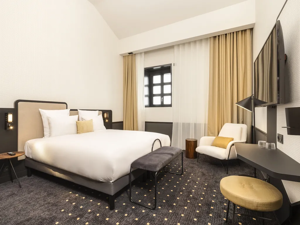 Modern hotel room featuring a white king-size bed, cream-colored armchair, black desk, window with beige curtains, and wall-mounted television with contemporary black and gold furnishings.