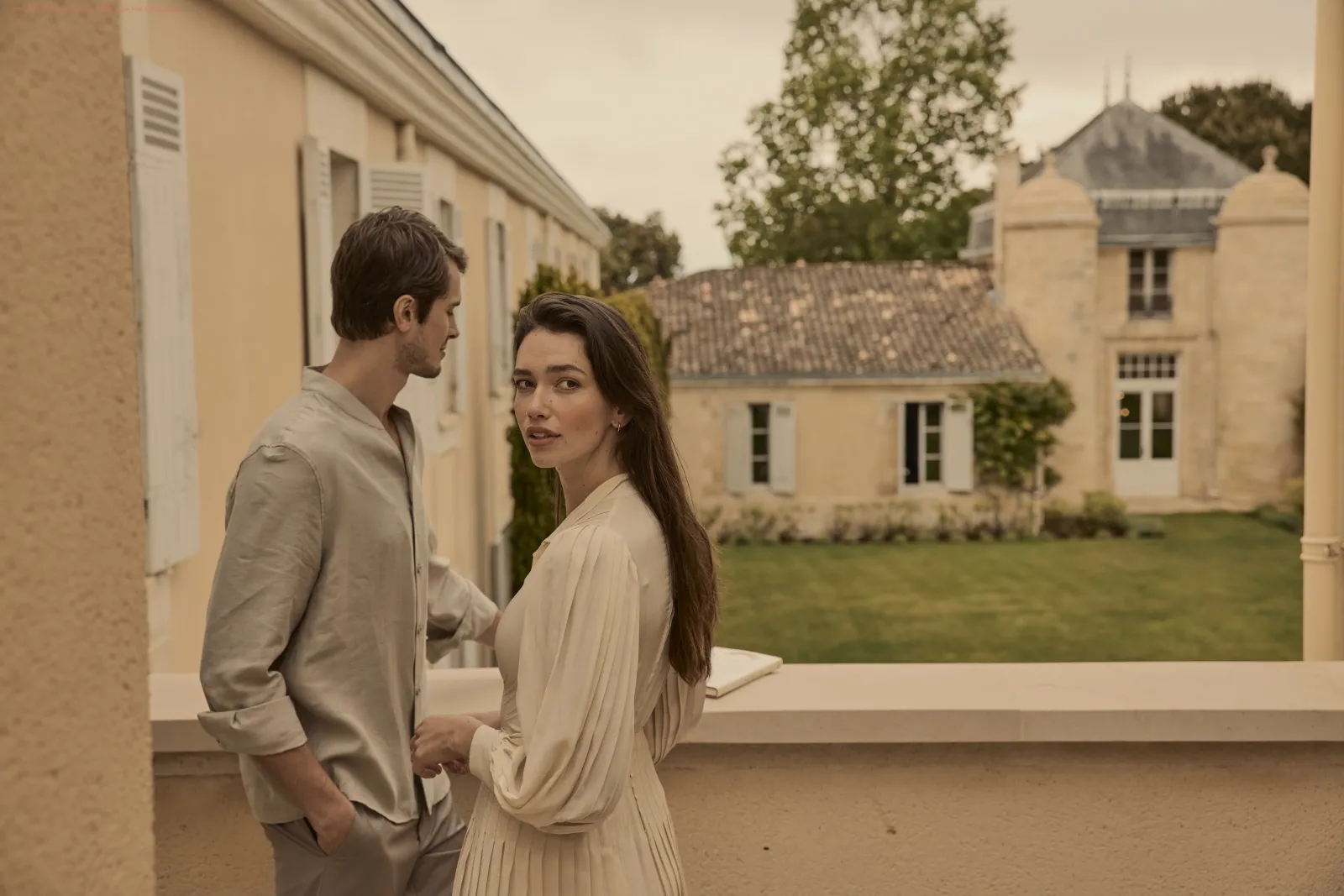 Two people in elegant attire converse on a terrace overlooking stone buildings in Bordeaux style architecture and a green garden.