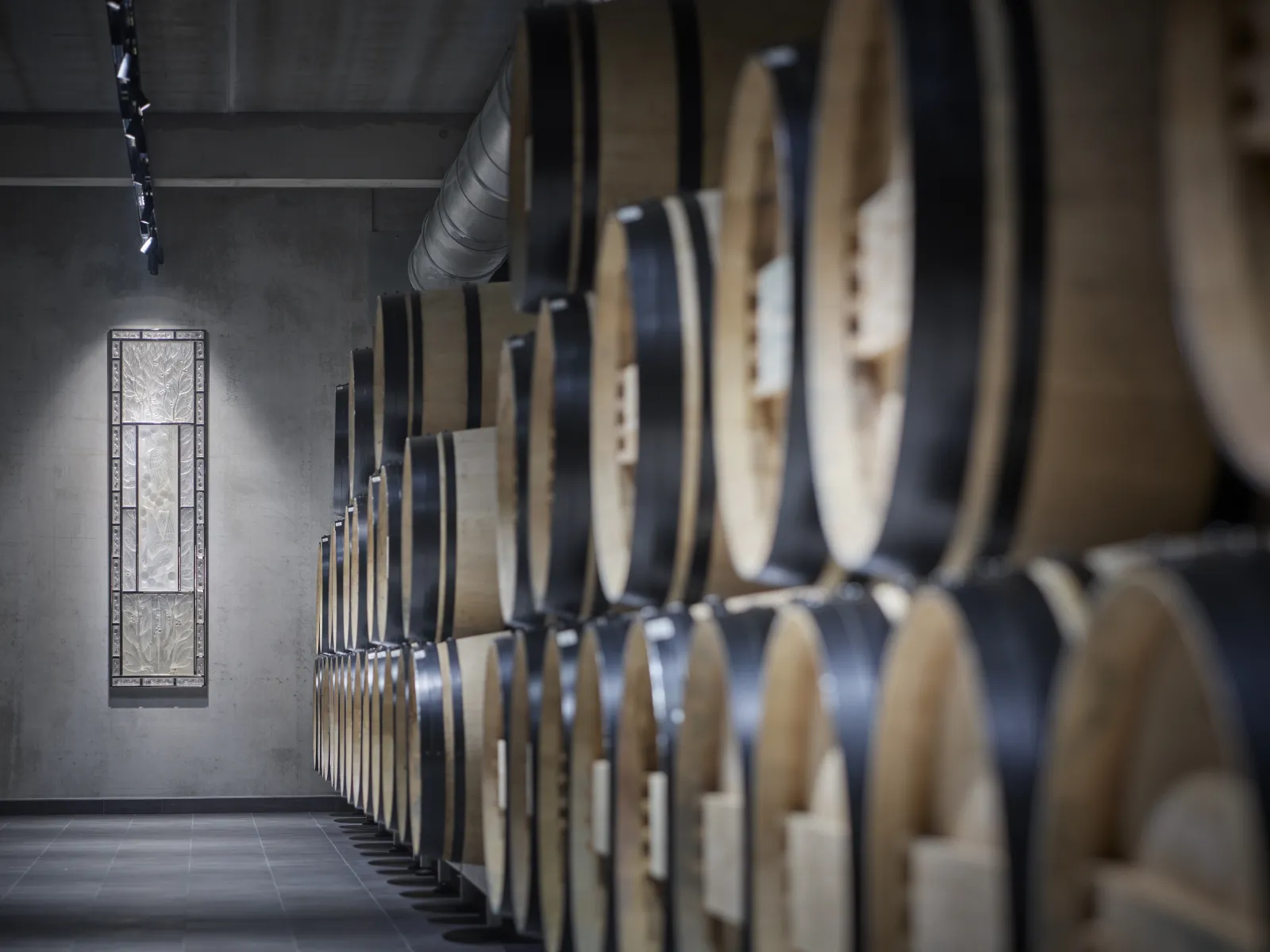 Row of oak barrels in the cellar of Château Peby Faugères in Bordeaux, with a narrow window and ventilation system visible